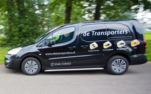 Zwarte bestelauto van De Transporters rijdt over een weg, met bedrijfslogo en diensten vermeld op de zijkant.