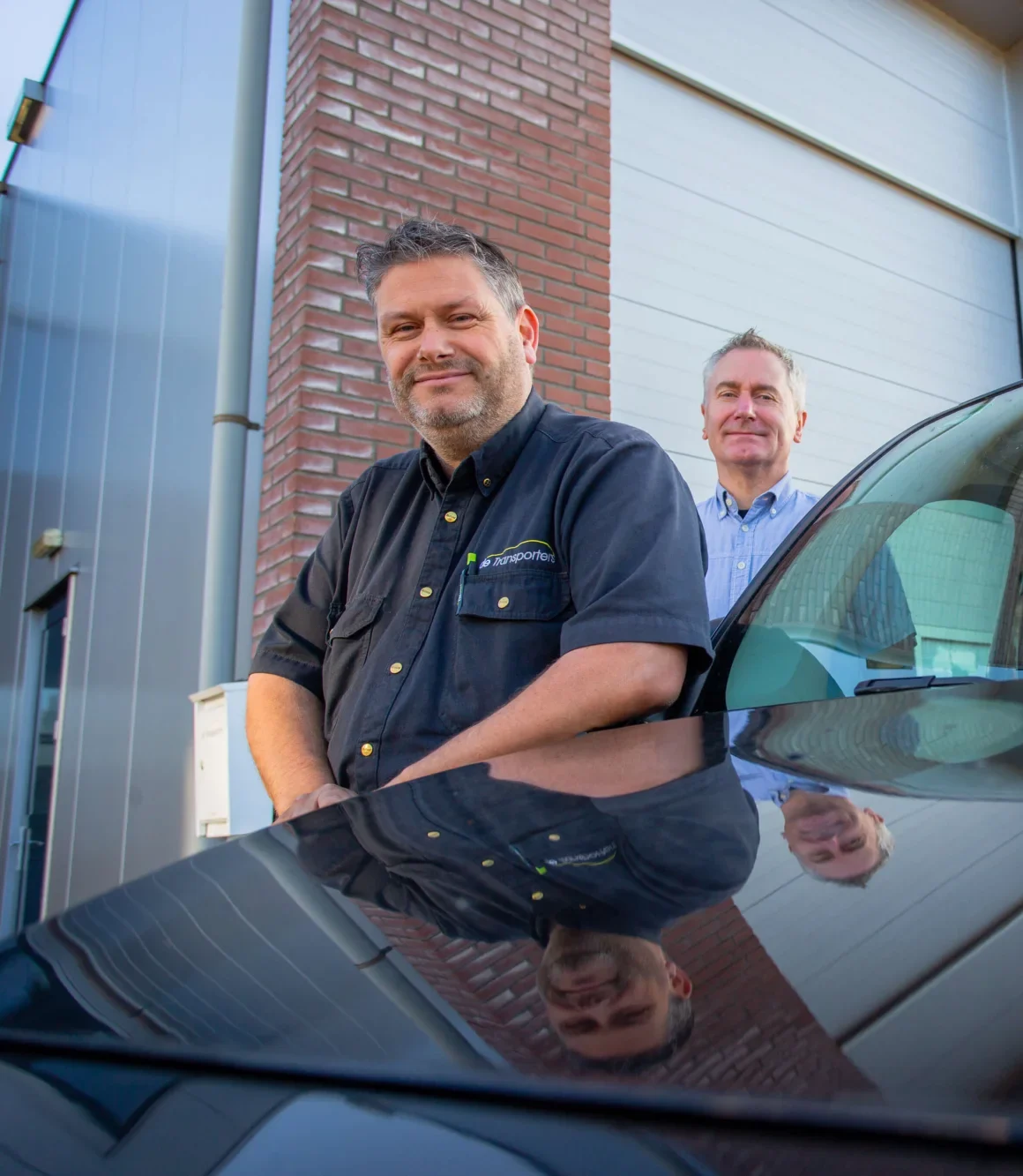 Twee mannen bij een bedrijfsgebouw, waarvan één in werkkleding van De Transporters leunend op een auto, met hun reflectie zichtbaar in de motorkap.