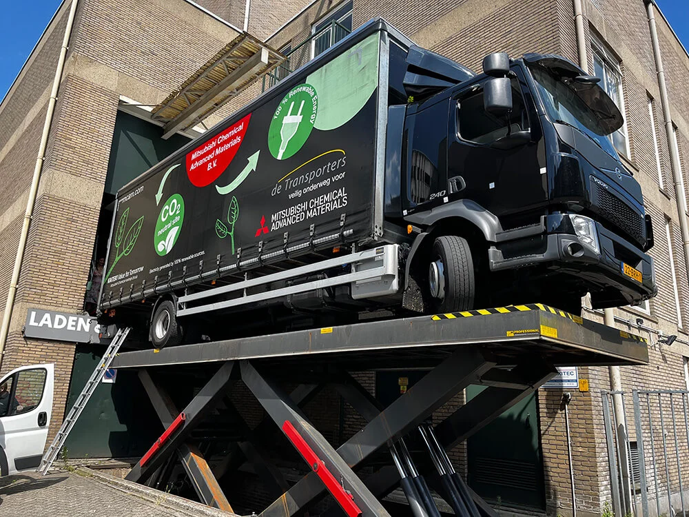 Vrachtwagen van De Transporters op een hydraulisch hefsysteem bij een laadperron van Mitsubishi Chemical Advanced Materials, met duurzame branding op de zijkant.