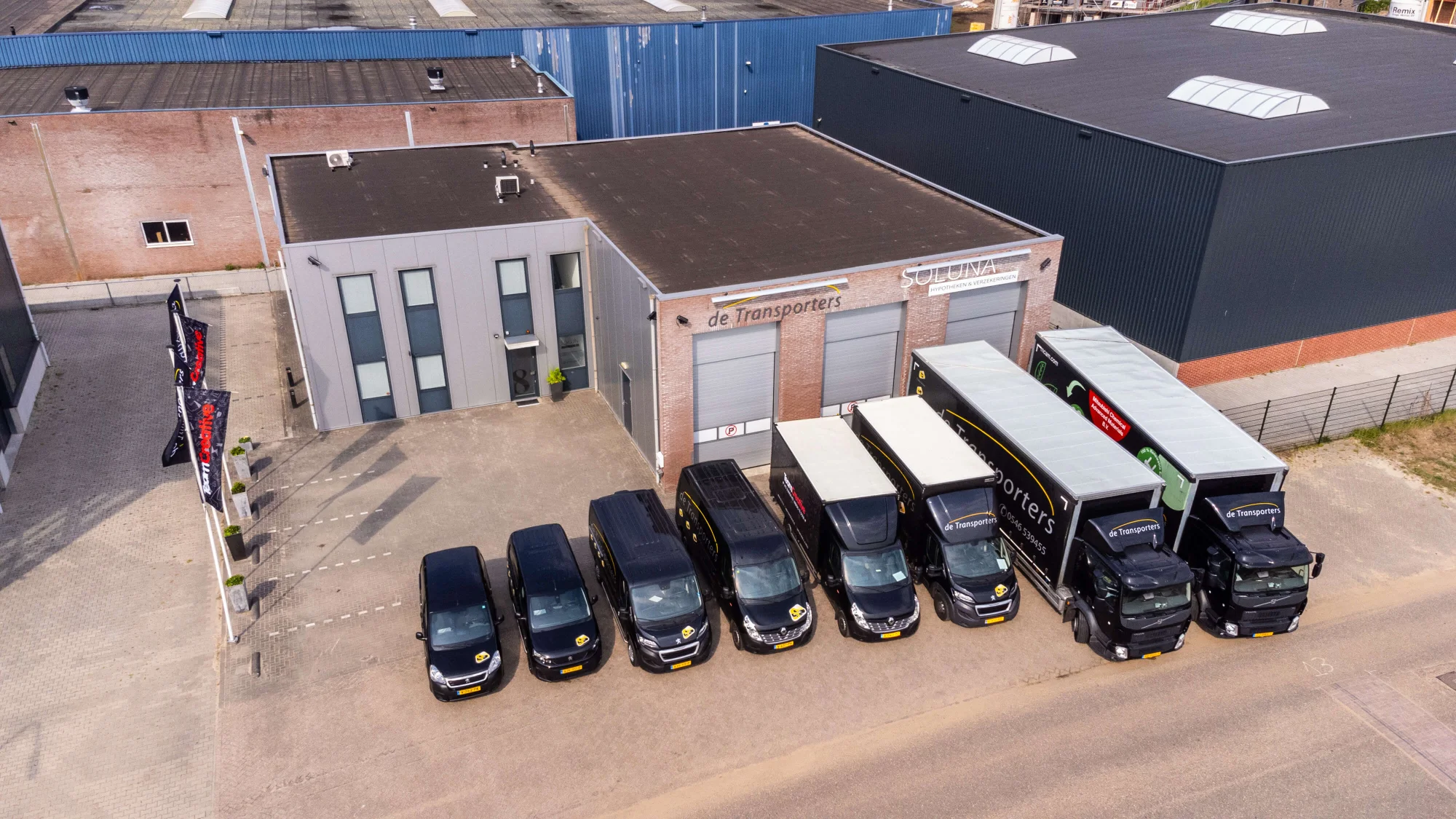 Luchtfoto van het pand van De Transporters in Almelo, met meerdere bestelbussen en vrachtwagens geparkeerd voor de gevel en vlaggen bij de entree.