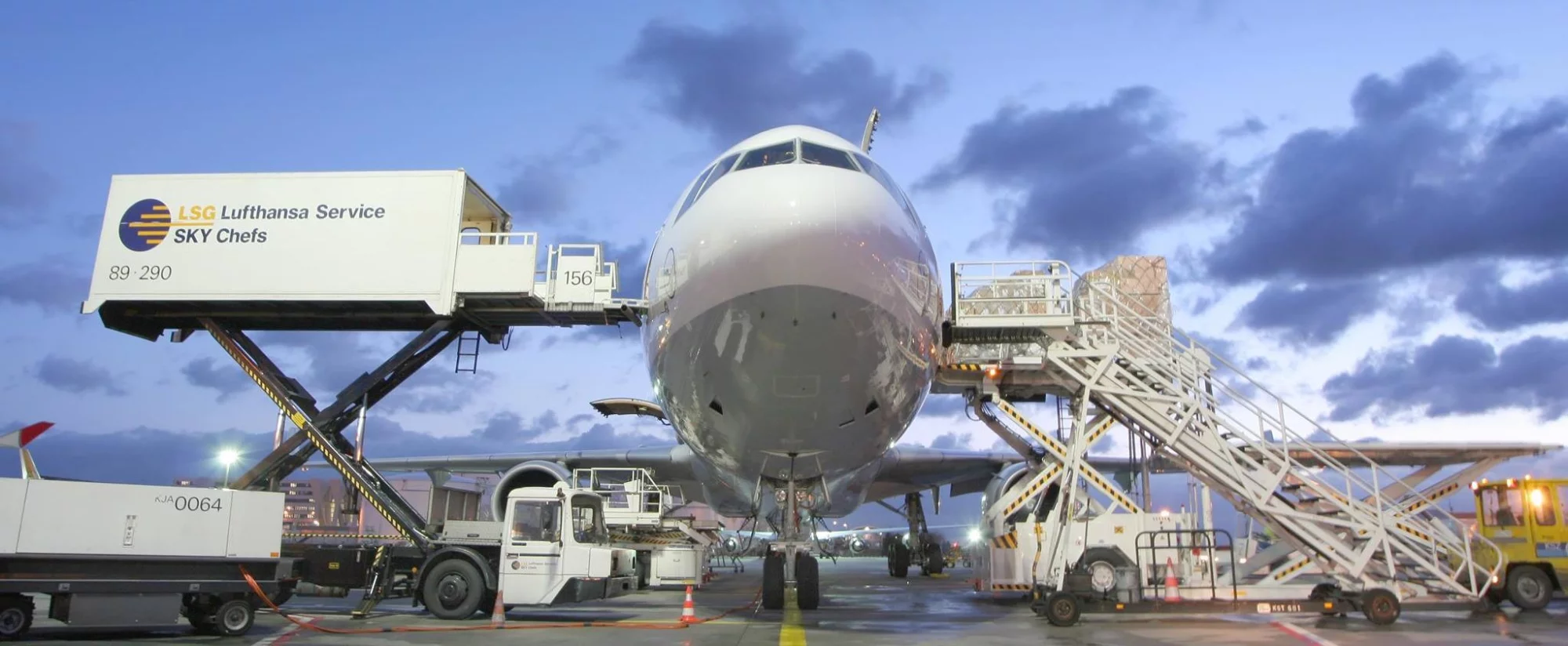Vliegtuig aan de grond op een luchthaven tijdens bevoorrading en belading, met cateringtruck van LSG Sky Chefs en een vrachtplatform met goederencontainers aan weerszijden van de neus.
