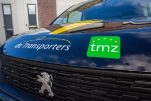 Close-up van de motorkap van een Peugeot-bestelwagen met logo’s van De Transporters en TMZ-keurmerk met vijf sterren op een groene achtergrond.