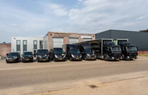 Vooraanzicht van het pand van De Transporters met een complete rij bedrijfsvoertuigen, waaronder bestelbussen en vrachtwagens, geparkeerd op een industrieterrein.