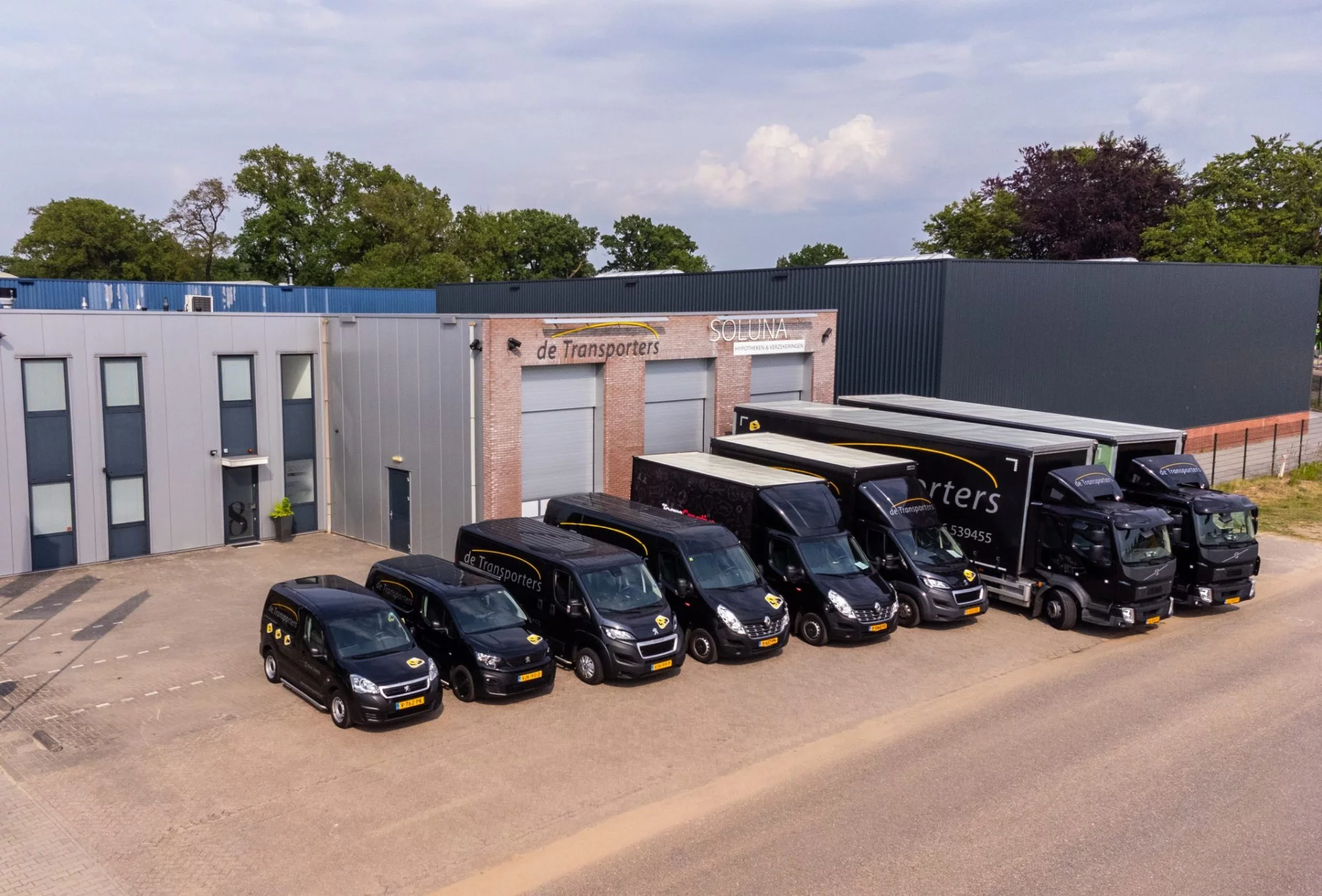 Luchtfoto van het bedrijfspand van De Transporters met een complete vloot van bestelbussen en vrachtwagens, geparkeerd op de oprit naast het kantoor.