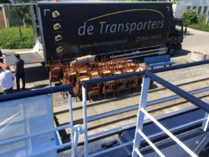 Vrachtwagen van De Transporters staat open met een grote lading gestapelde stoelen op de stoep, terwijl enkele personen erbij staan op een zonnige dag.