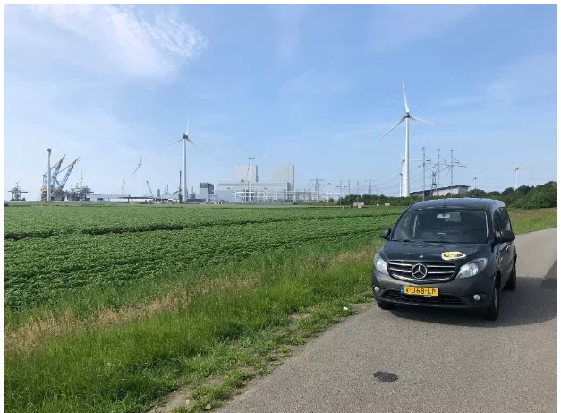 Zwarte bestelwagen van De Transporters met gele sticker staat geparkeerd langs een landweg met op de achtergrond windmolens, industrie en een veld met groene gewassen.