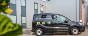 Bestelbus van De Transporters op een bedrijventerrein, met zichtbare diensticonen en contactgegevens op de zijkant.