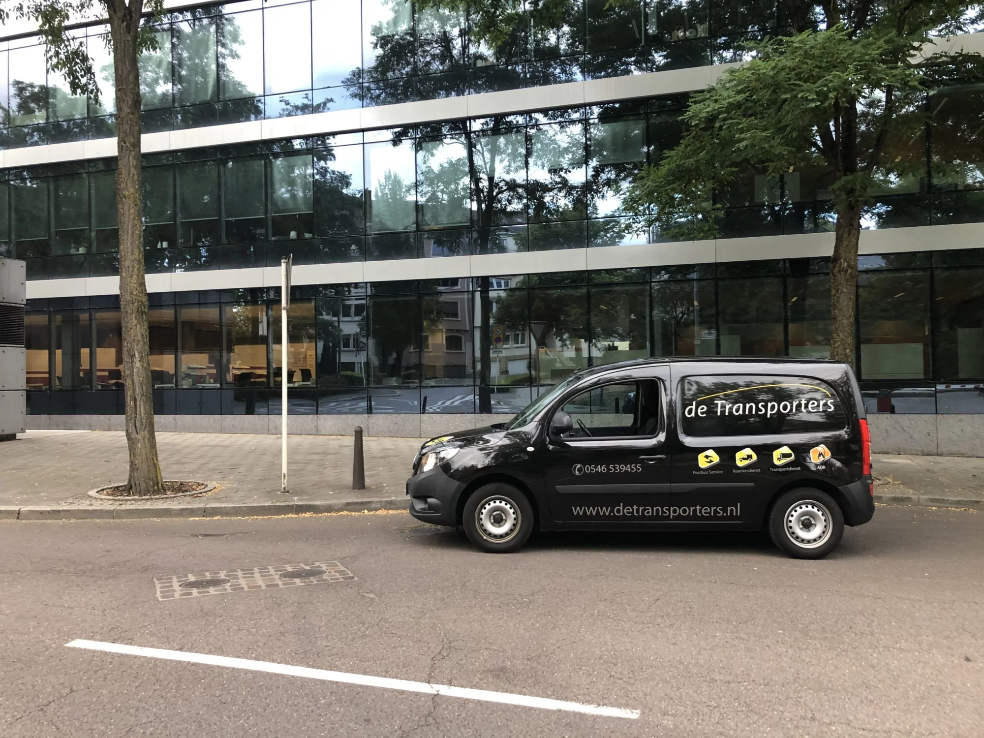 Bestelbus van De Transporters geparkeerd voor een kantoorpand, met bedrijfslogo en website-adres op de zijkant