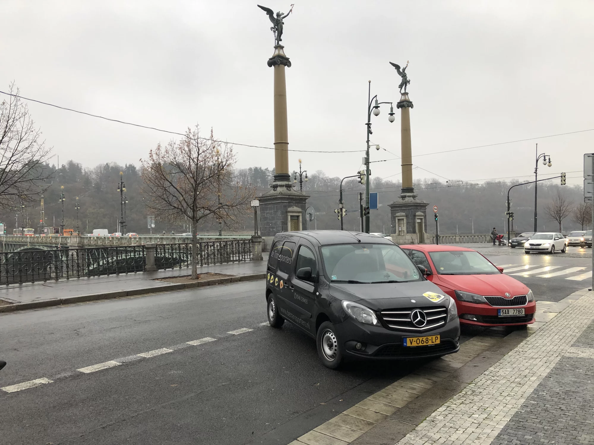 Bestelbus van De Transporters geparkeerd naast een rode auto bij een klassieke brug met standbeelden in een mistige, stedelijke omgeving.