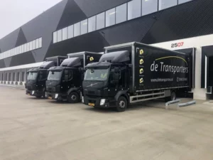 Drie zwarte vrachtwagens van de Transporters geparkeerd bij een modern distributiecentrum, klaar voor transport.