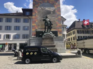 Zwarte bestelbus van De Transporters geparkeerd voor een standbeeld van Wilhelm Tell op een plein in een stad met typische Europese architectuur en een Zwitserse vlag zichtbaar.