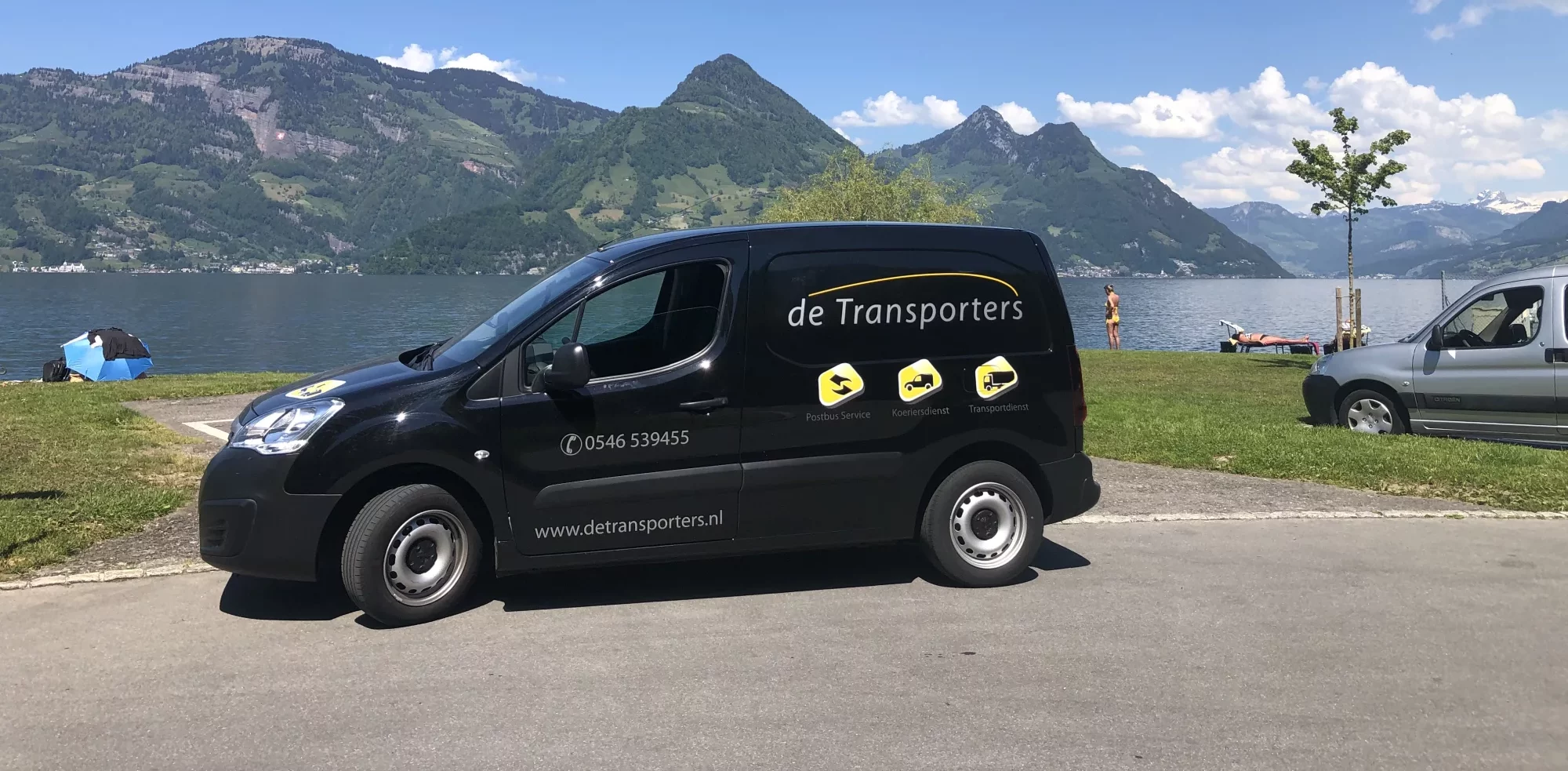 Zwarte bestelbus van De Transporters geparkeerd aan het water met een bergachtig landschap op de achtergrond en mensen die aan het recreëren zijn bij het meer.