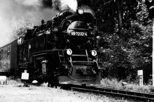 Zwart-witfoto van een rijdende stoomlocomotief met nummer 99 7232-4, omringd door bomen en rookpluimen die opstijgen uit de schoorsteen.