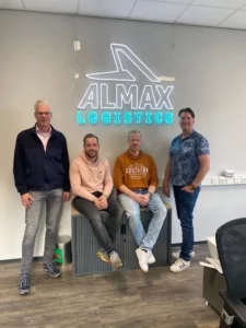 Groepsfoto van vier mannen voor een wand met het verlichte logo van ALMAX LOGISTICS; twee mannen staan en twee zitten op een grijze kast, allen lachend in een kantooromgeving.