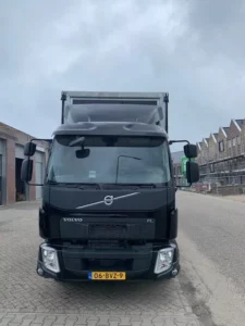 Vooraanzicht van een zwarte Volvo FL vrachtwagen met Nederlands kenteken, geparkeerd op een bedrijventerrein met bouwactiviteiten op de achtergrond.