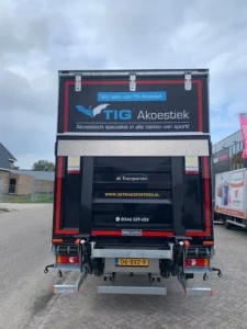 Achterzijde van een vrachtwagen van De Transporters met reclame voor TIG Akoestiek, gefotografeerd op een industrieterrein. Teksten en logo’s van beide bedrijven zijn zichtbaar, evenals het kenteken en de laadklep van de truck.