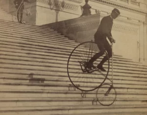Man op een hoge bi (antieke fiets met groot voorwiel) rijdt een brede stenen trap af voor een statig gebouw.